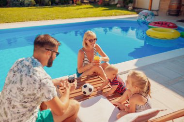 Genç ve güzel bir aile, güneşli bir yaz gününü yüzme havuzunun yanında geçirir ebeveynler dinlenir ve kokteyl içerken çocuklar da yanlarında güneşlenir.