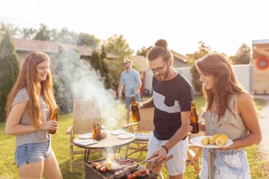 Bir grup neşeli genç arkadaş arka bahçedeki barbekü partisinde eğleniyor, et pişiriyor, bira içiyor, badminton oynuyor ve güneşli bir yaz gününde dışarıda dinleniyor.