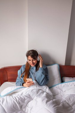 Güzel genç bir kadın yatakta oturuyor, sabahları evde dinleniyor, kulaklık takıyor ve akıllı telefon kullanarak internette sörf yapıyor.