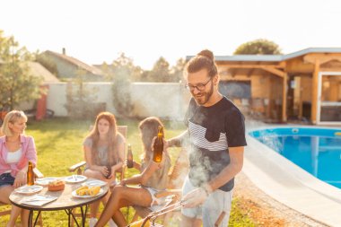Bir grup genç arkadaş yüzme havuzunun yanında barbekü partisi veriyor erkekler ön planda et pişiriyor ve bira içiyor kadınlar arka planda oturup dinleniyor.