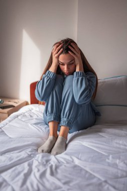 Endişeli ve çaresiz genç bir kadın yatakta kıvrılmış, el ele tutuşmuş, ciddi ve düşünceli.