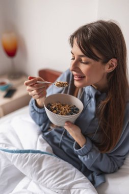 Beautiful young woman wearing pajamas sitting in bed in the morning, having cereal with fresh berries for breakfast and enjoying leisure time at home clipart