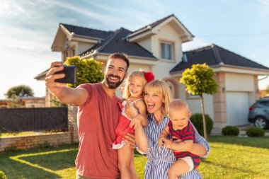 Güzel mutlu aile yeni evlerinin önünde akıllı telefonları kullanarak selfie çekerken eğleniyor.