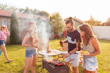 Bir grup neşeli genç arkadaş arka bahçede barbekü partisi veriyor, et pişiriyor, bira içiyor, badminton oynuyor ve güneşli bir yaz gününde dışarıda dinleniyor.