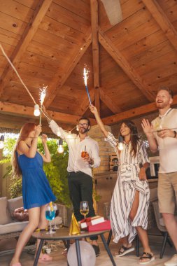 Bir grup neşeli genç arkadaş havai fişeklerle el sallıyor, doğum günü partisinde dans ediyor ve kokteyl içiyor.