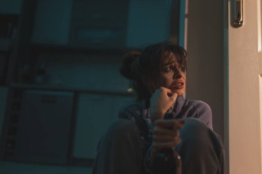 Üzgün sarhoş bir kadın karanlıkta yerde makyajla oturmuş şişeden şarap içiyor ve ağlıyor.