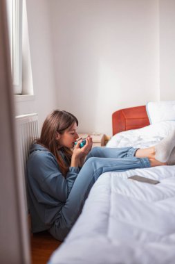 Pijama giyen güzel genç bir kadın yatak odasındaki yatağın yanında oturuyor, sabah kahvesini içiyor ve evde dinleniyor.
