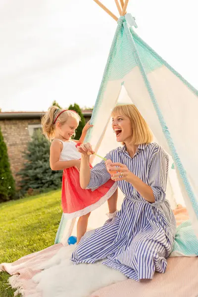 美丽的母亲和小女儿在后院露营和玩耍 坐在帐篷里 制造和捕捉肥皂泡 在阳光明媚的夏日在户外放松 — 图库照片