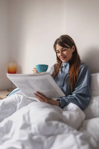 Beautiful young woman wearing pajamas relaxing in bed after waking up, reading newspaper and drinking her morning coffee