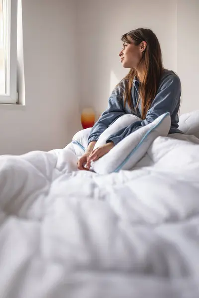 Beautiful young woman wearing pajamas holding a pillow and sitting in bed in the morning, looking through the window pensive and absent