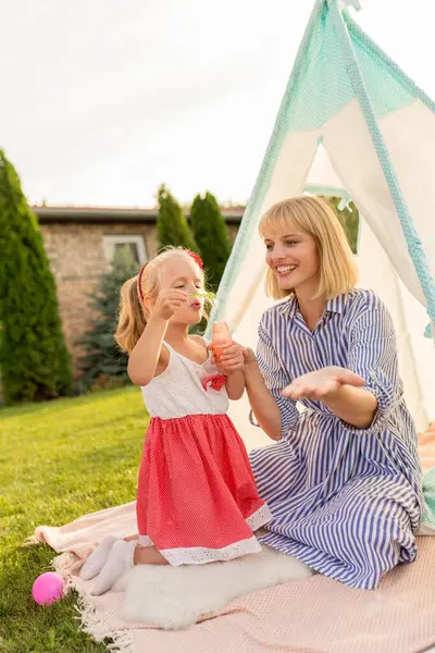 美丽的母亲和小女儿在后院露营和玩耍 坐在帐篷里 吹着肥皂泡 在阳光明媚的夏日在户外放松 — 图库照片