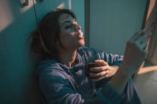 暗闇の中で床に座り ワインを飲んでタバコを吸い 動揺して絶望する抑圧された若い女性 — ストック写真