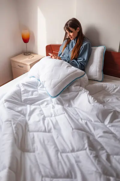 漂亮的年轻女人早上醒来后 穿着睡衣 在床上用智能手机 — 图库照片