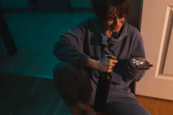 暗い酔っ払いのキッチンフロアに座り ワインのボトルと一握りの錠剤を保持する絶望的な若い女性 物質乱用 うつ病 自殺の概念 — ストック写真