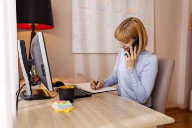 Kadın müşteriyle telefon görüşmesi yapıyor ve ev ofisinde çalışırken planlama yapıyor.