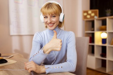 Evdeki ofisinde masasında oturan, kulaklık takan ve uzaktan çalışırken parmağını kaldıran bir kadın.