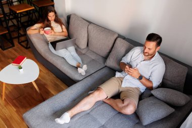 Bir çift evde boş vakitlerini birlikte geçiriyorlar, oturma odasındaki kanepede dizüstü bilgisayar ve akıllı telefon kullanarak yatıyorlar, online haberler okuyorlar.