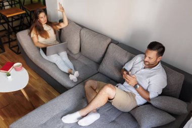 Yüksek açılı bir bakış açısıyla, çift boş zamanlarını evde birlikte geçiriyorlar, oturma odasındaki kanepede dizüstü bilgisayar ve akıllı telefon kullanarak uzanıyorlar, internet haberleri okuyorlar.