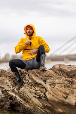 Düşmüş bir ağaç gövdesinde oturan bir adam, bulutlu bir sonbahar gününde nehir kenarında koşarken, mesafeye dalgın dalgın bakıyor.