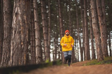 Kulaklık takan, müzik dinleyen ve bulutlu bir sonbahar gününde parkta koşan aktif bir adam.