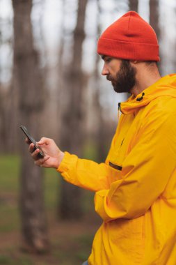 Bir adam bulutlu bir sonbahar gününde parkta koşarken, bir ağaca yaslanır ve akıllı bir telefon kullanır.
