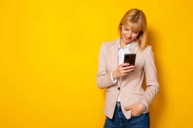 Güzel, kendine güvenen genç bir iş kadını sarı arkaplanda izole edilmiş ve kopyalama alanı olan akıllı telefon kullanarak kısa mesaj yazıyor.