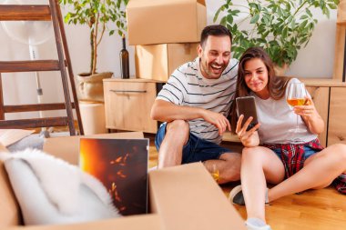 Genç çift yeni bir daireye taşınırken akıllı telefon kullanıp şarap içiyorlar.