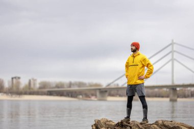 Düşmüş bir ağaç gövdesinde duran atletik adam, bulutlu bir sonbahar gününde nehir kenarında koşarken dinleniyor ve dinleniyor.