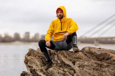 Düşmüş bir ağaç gövdesinde oturan bir adam, bulutlu bir sonbahar gününde nehir kenarında koşarken, mesafeye dalgın dalgın bakıyor.