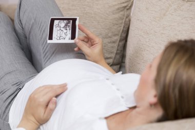 Hamile bir kadın, dairesinin oturma odasında bir kanepede uzanıp bebeğinin ultrason fotoğrafına bakıyor.