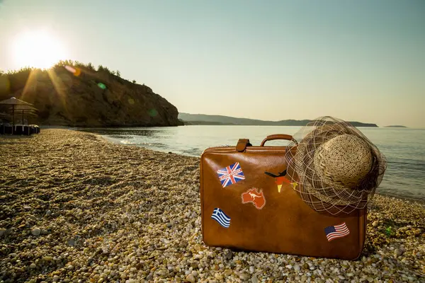 Vintage suitcase with flag stickers and a straw hat, placed on the beach with peacefull sea during sunrise on Thassos island in Greece
