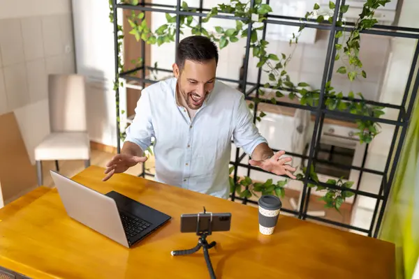 ホームオフィスで働く男 三脚に設定されたラップトップコンピュータとスマートフォンを使って彼のビデオを録画する — ストック写真