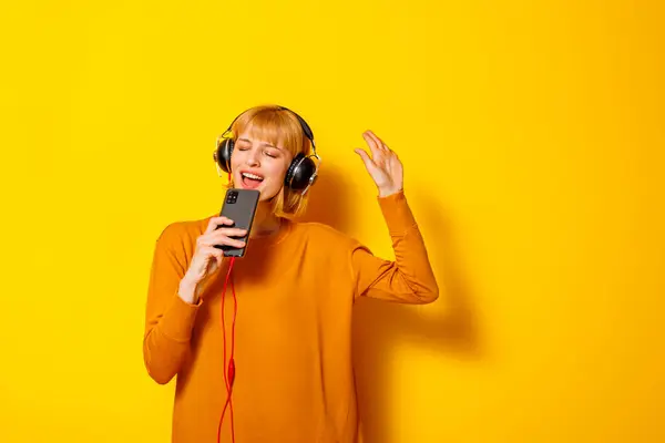コピースペースで黄色の背景に隔離されたマイクとして音楽やスマートフォンを使用して歌う楽しいヘッドセットを身に着けている美しい若い女性 — ストック写真