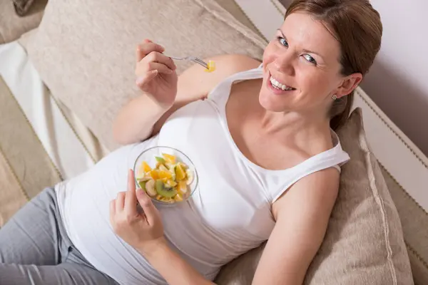 孕妇坐在客厅的沙发上 吃着水果沙拉 — 图库照片