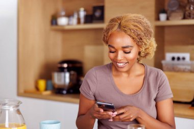 Güzel genç bir kadın mutfak tezgahında oturmuş kahvaltı yapıyor ve internette akıllı telefon kullanarak sörf yapıyor, sabahları evde dinleniyor.