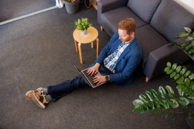 Ofis zemininde oturan, kanepeye yaslanan ve dizüstü bilgisayarla çalışan erkek yazılım geliştiricisinin yüksek açılı görüntüsü