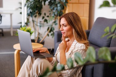 Serbest çalışan bir kadın, yerde oturuyor kanepeye yaslanıyor ve ev ofisinde dizüstü bilgisayarla çalışıyor.