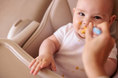 Anne, bebeği kaşıkla besliyor, ilk katı yiyeceği sunuyor, bebek yüksek sandalyede oturuyor ve lapayla oturmuş, gülümsüyor ve yiyor.