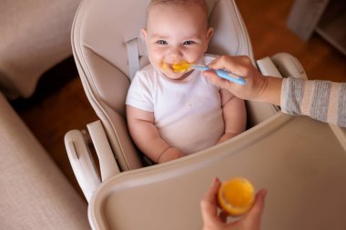 Annesinin neşeli erkek çocuğunu lapayla beslediği yüksek açılı manzara, bebek sandalyede oturuyor ve gülümsüyor ve yemek yiyor.
