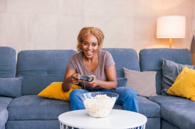 Güzel, genç Afrikalı bir kadın oturma odasındaki kanepede oturmuş evde oyun oynayarak eğleniyor.