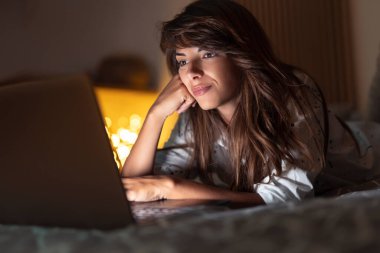 Güzel bir kadın gece geç saatlerde evde dinleniyor, yatakta uzanıyor ve dizüstü bilgisayarla internette sörf yapıyor.