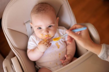 Annesinin neşeli erkek çocuğunu lapayla beslediği yüksek açılı manzara, bebek sandalyede oturuyor ve gülümsüyor ve yemek yiyor.