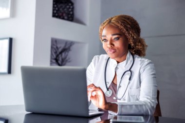 Ofisindeki masasında oturan kadın doktor, hasta ile çevrimiçi video görüşmesi yaparken dizüstü bilgisayarla tıbbi rapor yazıyor.