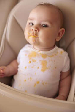 Sevimli küçük oğlan yüksek sandalyede oturmuş lapaya bulanmış, komik suratlar yapıyor ve yemek yemeyi reddediyor.