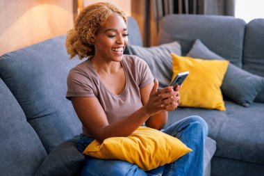 Güzel genç bir kadın evde dinleniyor, oturma odasındaki kanepede oturuyor ve akıllı telefonuyla video görüşmesi yaparken gülümsüyor.