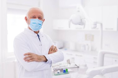 Hastanede kollarını kavuşturmuş, tıbbi koruyucu maske takan kıdemli erkek doktorun portresi.