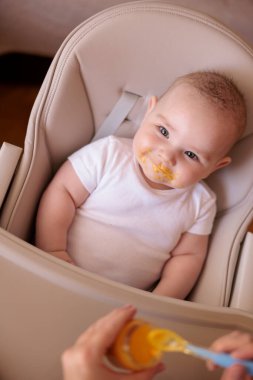 Annesinin neşeli erkek çocuğunu lapayla beslediği yüksek açılı manzara, bebek sandalyede oturuyor ve gülümsüyor ve yemek yiyor.