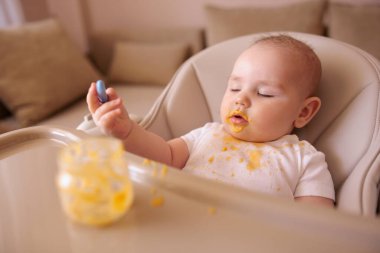 Neşeli komik çocuk, elinde kaşıkla yüksek sandalyede oturmuş, kabak lapası yerken kahkahalarla gülüyor.