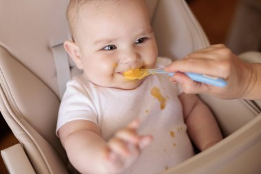Annesinin neşeli erkek çocuğunu lapayla beslediği yüksek açılı manzara, bebek sandalyede oturuyor ve gülümsüyor ve yemek yiyor.