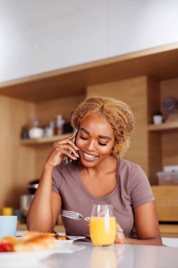 Güzel genç bir kadın mutfak tezgahında oturmuş kahvaltı yapıyor ve telefonda konuşuyor, sabahları evde dinleniyor.